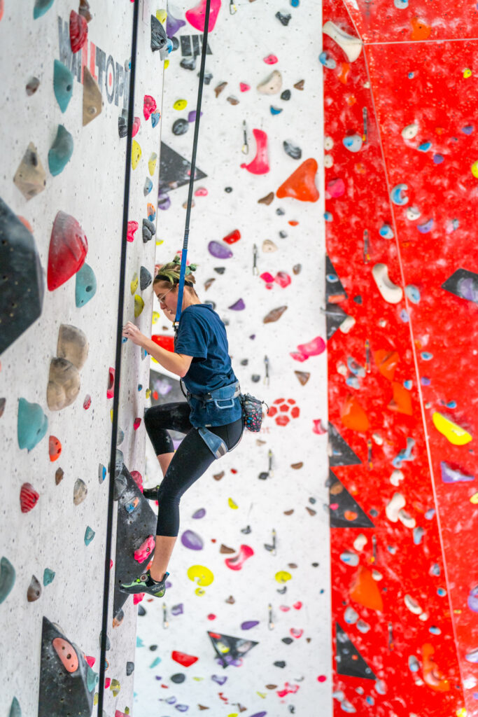 Woman climbing a vertical indoor wall at Redpoint Bristol, using an auto belay system and stepping between colourful holds beside a large red overhang section.
