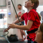 Young girl in a red top focuses on handling a climbing rope at Redpoint Bristol, looking up towards the wall while practising belaying techniques indoors.