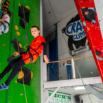 Boy in bright orange top descends an indoor climbing wall on a rope at Redpoint Bristol, with other climbers and colourful holds visible in the background.