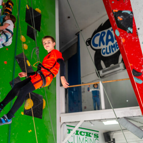 Boy in bright orange top descends an indoor climbing wall on a rope at Redpoint Bristol, with other climbers and colourful holds visible in the background.