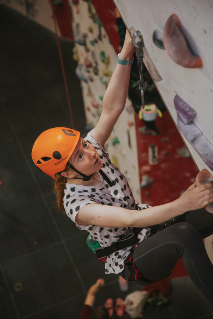 Close-up of a young climber in an orange helmet clipping into a quickdraw while looking up and gripping holds on an indoor climbing wall at Redpoint Bristol