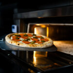 A freshly prepared pizza topped with pepperoni and green peppers sits on a metal peel as it’s slid into a hot oven, with warm light illuminating the crust and bubbling cheese.