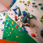 A climber stretches across an overhanging wall with large volumes and colourful holds, while others observe and prepare on the padded floor at Redpoint Climbing Centre in Bristol.