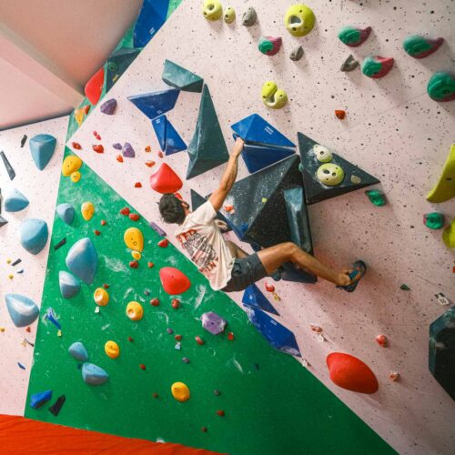 A climber stretches across an overhanging wall with large volumes and colourful holds, while others observe and prepare on the padded floor at Redpoint Climbing Centre in Bristol.