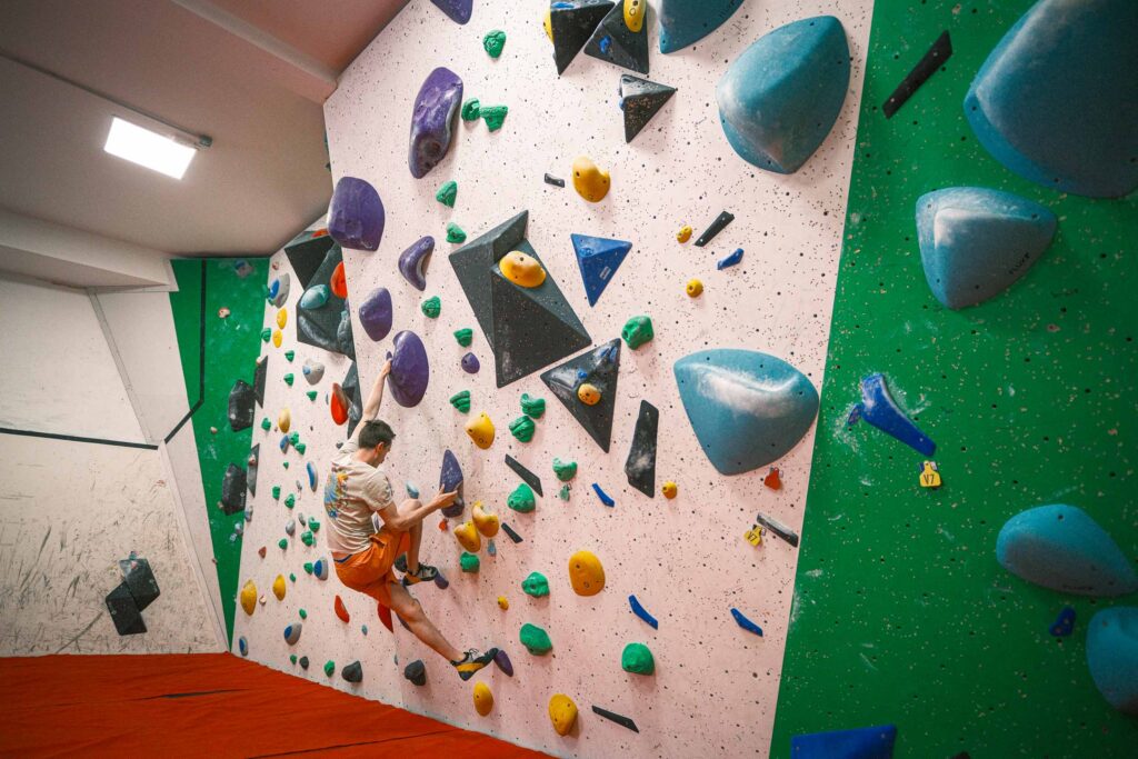 A climber works across a colourful indoor bouldering wall featuring large volumes and varied holds, set above padded flooring at Redpoint Climbing Centre in Bristol.