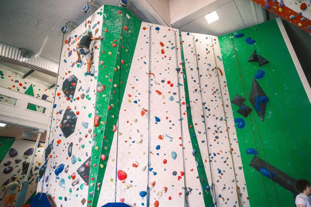 A climber makes their way up a high indoor wall with a variety of colourful holds and routes, showcasing the scale and variety of climbing options at Redpoint Climbing Centre in Bristol.