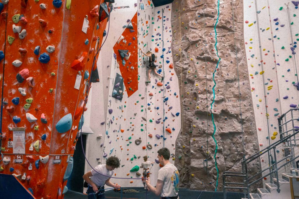 Wide view of climbers tackling different indoor walls, including a textured rock face and colourful routes, while others manage ropes and prepare climbs at Redpoint Climbing Centre in Bristol.