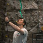 Male belayer holding the rope and looking up attentively, managing safety for a climber on a textured indoor rock wall with illuminated route line at Redpoint Climbing Centre in Bristol.