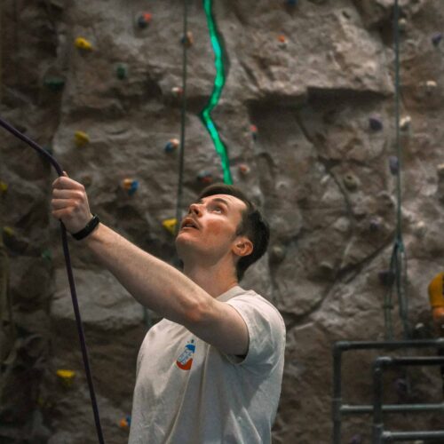 Male belayer holding the rope and looking up attentively, managing safety for a climber on a textured indoor rock wall with illuminated route line at Redpoint Climbing Centre in Bristol.