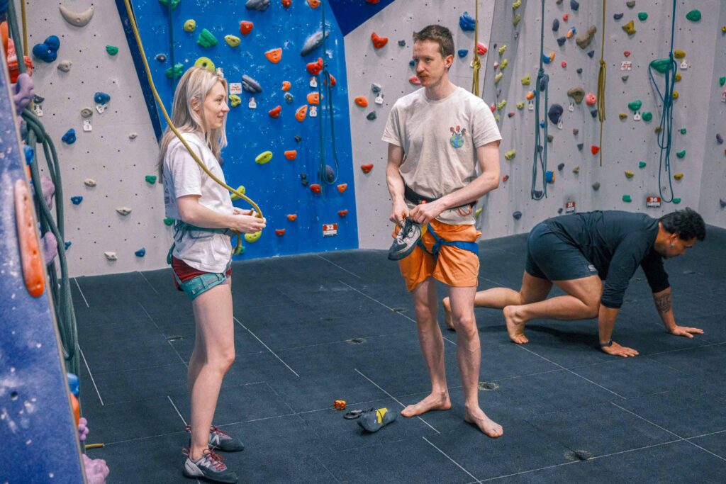 People getting ready to climb, adjusting harnesses and shoes on the mat area beneath colourful indoor climbing walls at Redpoint Bristol.