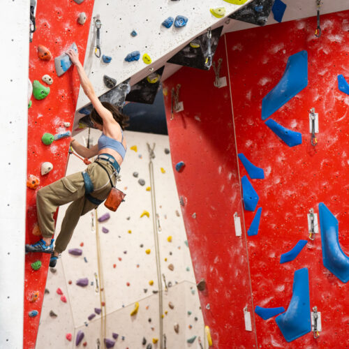 A woman lead climbs a steep red panel, reaching high for a hold while clipped into the rope, with the large red feature wall and blue holds beside her.