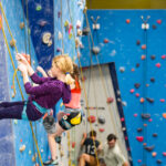 A young girl in a harness leans back on a rope on the blue training wall, with other climbers and belayers in the background.