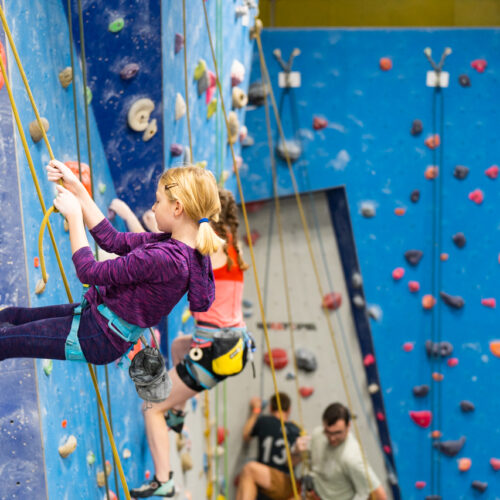 A young girl in a harness leans back on a rope on the blue training wall, with other climbers and belayers in the background.