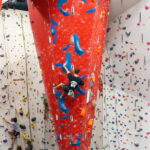 A man climbs a roped route on the tall red feature wall with large blue holds, while other climbers belay and practise on nearby walls below.