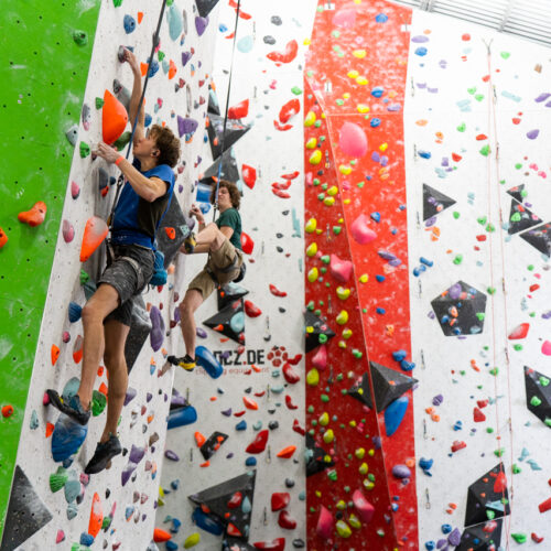 Two men climb separate roped routes on steep indoor walls, one on the green panel and one on the white wall with volumes and colourful holds.