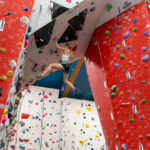 A woman lead climbs across a steep overhanging wall between red panels, reaching for the next hold while clipped into the rope.