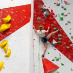 A woman lead climbs a red-and-white indoor wall, high-stepping onto a hold while clipped into the rope with quickdraws above and below.