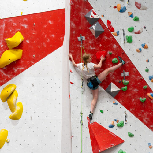 A woman lead climbs a red-and-white indoor wall, high-stepping onto a hold while clipped into the rope with quickdraws above and below.