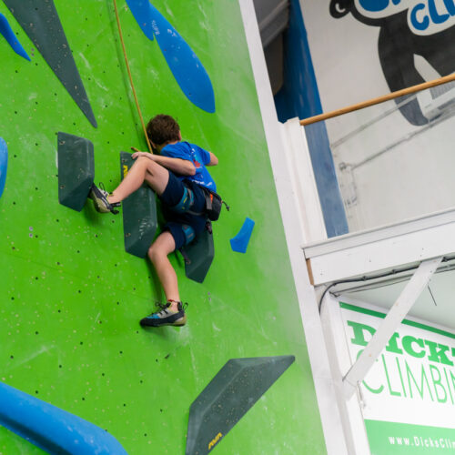 A boy climbs across a steep green training wall on large blue and black holds, clipped into a rope and wearing a harness.