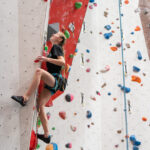A young girl climbs the lower section of a roped route, clipped into the rope and stepping onto holds beside a red overhanging panel.