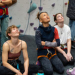 Three women sit on the padded floor in harnesses, looking up and following a climber on the wall above.